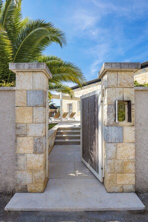 A gated entrance with stone pillars, a pathway leading to a covered seating area, and a palm tree.