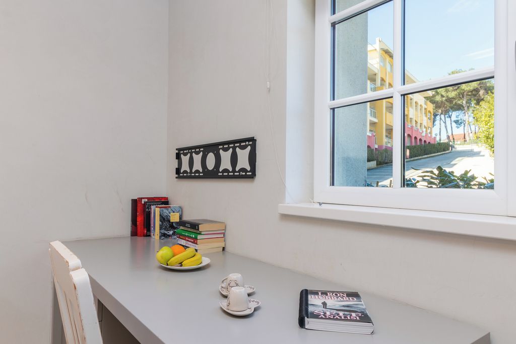 A room with a window, a table with books, a fruit bowl, and a decorative wall piece.