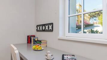 A room with a window, a table with books, a fruit bowl, and a decorative wall piece.