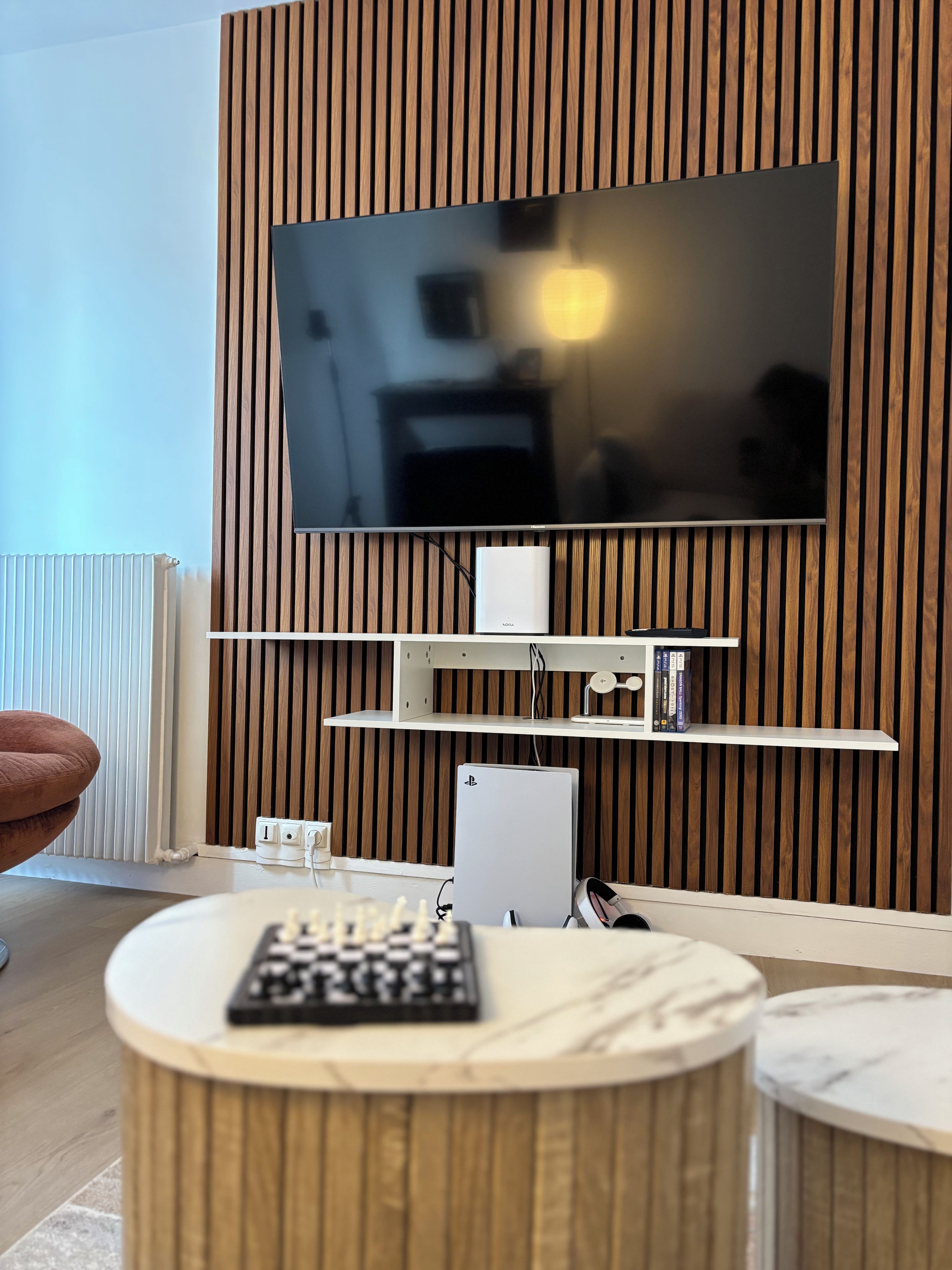 Un salon moderne avec un téléviseur à écran plat fixé sur un mur recouvert de panneaux en bois, une console blanche surmontée d’un jeu d’échecs et une console de jeux vidéo.
