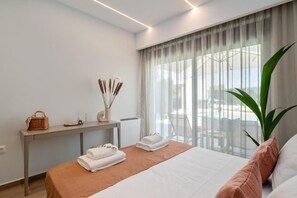 Ein modernes Hotelzimmer mit einem Bett, einem Nachttisch mit Korb und Kerzen, einem großen Fenster mit Vorhängen und einer Topfpflanze.