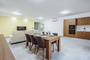 A modern dining area with a wooden table set for four, a sofa, and a television in the background. - Modern Maisonette With Pool in Gharghur (Hal Gharghur)