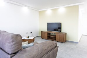 A living room with a flat-screen TV on a wooden stand, a grey sofa, and a small round table with a plant. - Spacious 4 Bedroom Apartment in Peaceful Gharghur (Hal Gharghur)