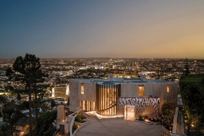 A modern building with a rooftop terrace, surrounded by trees and a cityscape at dusk. - Gatsby Mansion - Grand Hollywood Hills Estate with Endless City Vistas (Hollywood Hills, Los Angeles)