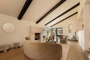 A spacious living room with a stone fireplace, wooden beams, and a large window offering a view of the outdoors. - Longridge Crest - Gated Studio City Canyon Home (Studio City)