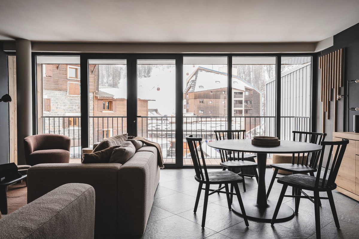A modern interior with a sofa, dining table, and chairs, large windows with a view of buildings and snow.