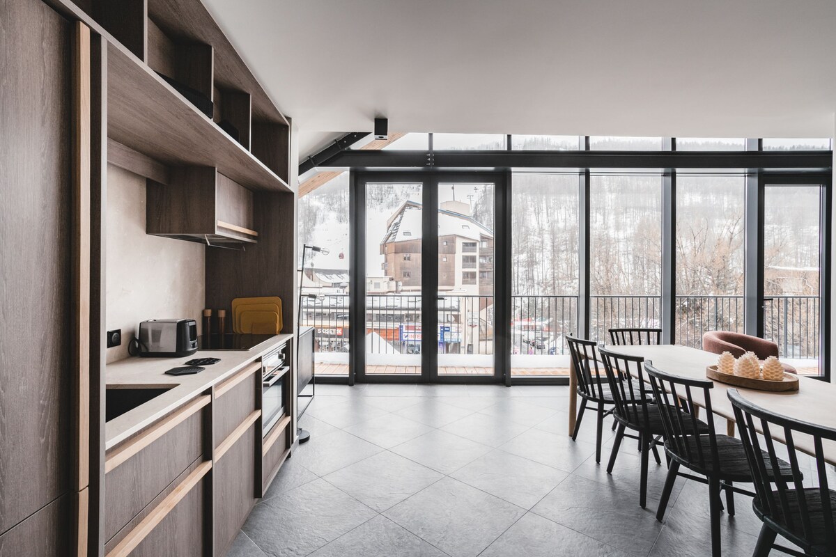 A modern kitchen with a dining area, large windows, and a view of a snowy landscape.