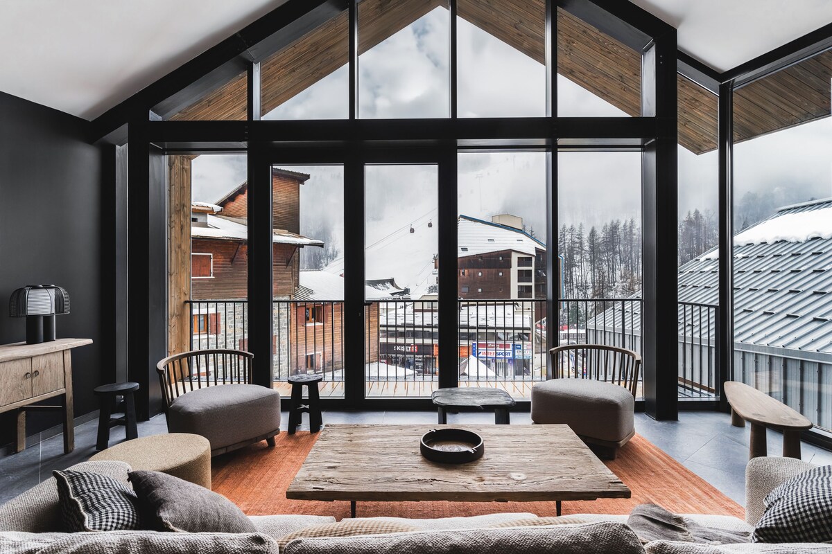 A spacious room with a large window overlooking a snowy mountain landscape, furnished with a wooden table, chairs, and a sofa.