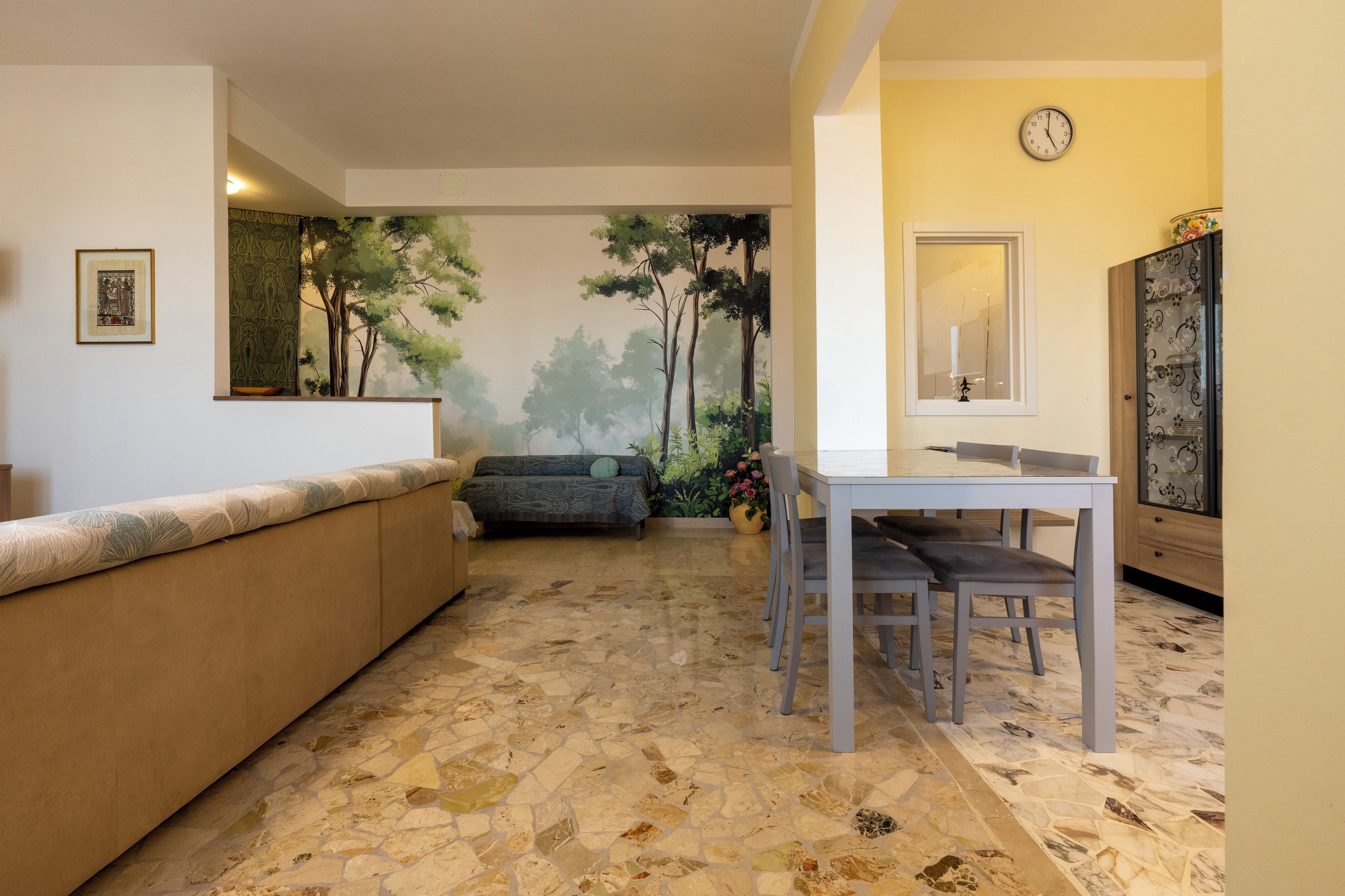 Una habitación con piso de baldosas de piedra, un sofá, una mesa de comedor con sillas, un reloj en la pared y un mueble con puerta estampada.