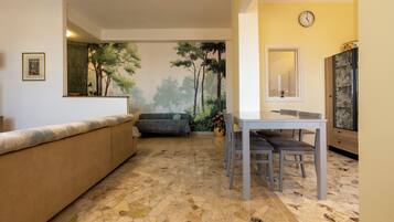 Una habitación con piso de baldosas de piedra, un sofá, una mesa de comedor con sillas, un reloj en la pared y un mueble con puerta estampada.