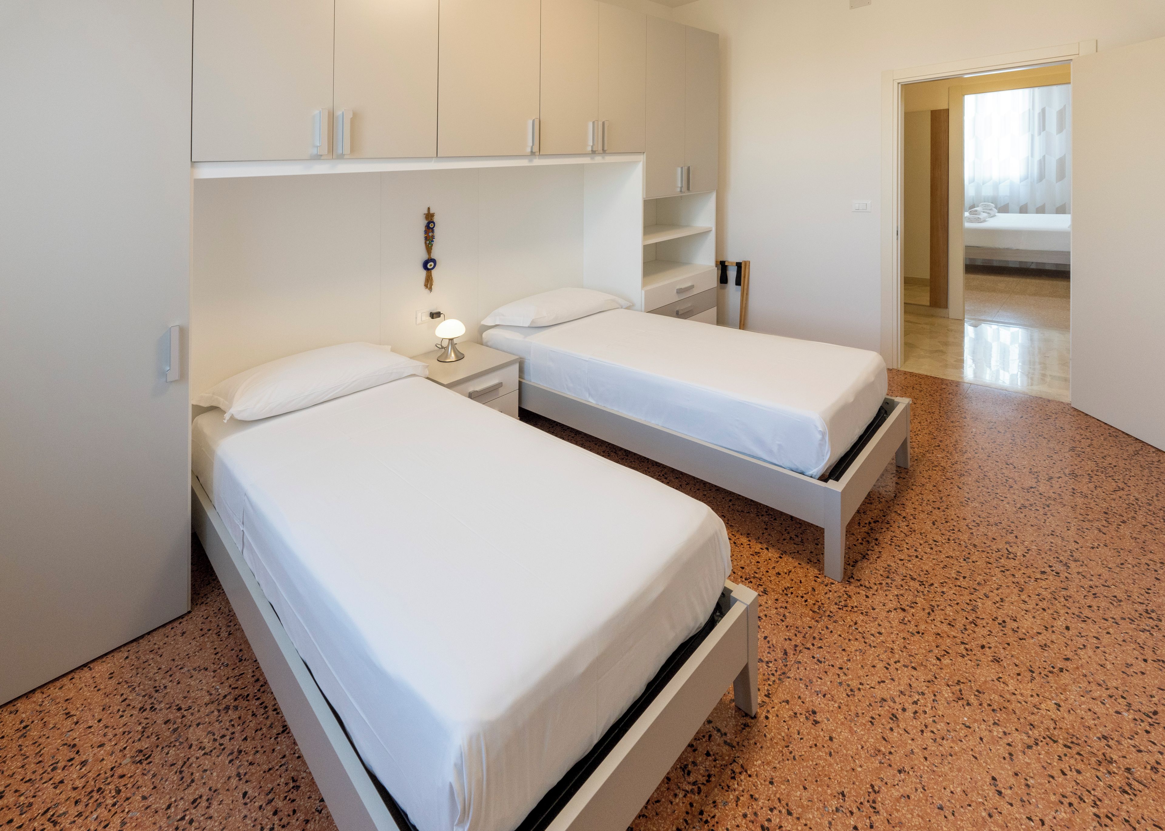 Een hotelkamer met twee enkele bedden, een kledingkast, een nachtkastje met een lamp en een deur naar een andere kamer.