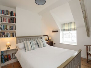 A bedroom with a large bed, a bookshelf, a window with curtains, and a wooden chair. - Swn y Mor (Beaumaris)