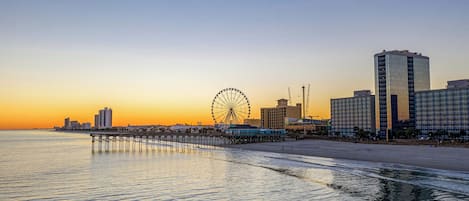 A coastal cityscape with a prominent Ferris wheel, modern high-rise buildings, and a pier extending into the water.