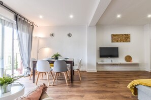 Un espace de vie moderne comprenant un coin repas, un téléviseur à écran plat fixé sur une étagère blanche et une grande fenêtre agrémentée de rideaux.