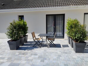En udendørs patio med et bord og stole, omgivet af plantede bede, og en bygning med et tag af fliser i baggrunden.