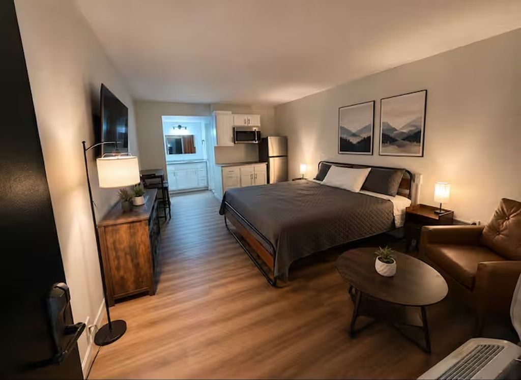 Habitación de hotel con cama, escritorio, silla, mesita, lámpara y microondas al fondo.