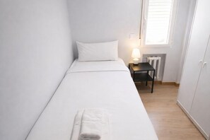Une petite chambre avec un lit simple, recouverte d’une courtepointe blanche, une table de chevet avec une lampe et un radiateur.