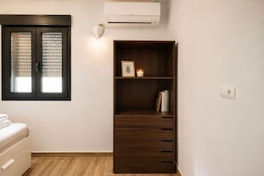 Een slaapkamer met een bed, een garderobe, een plank met boeken en een kaars, en een raam met jaloezieën.