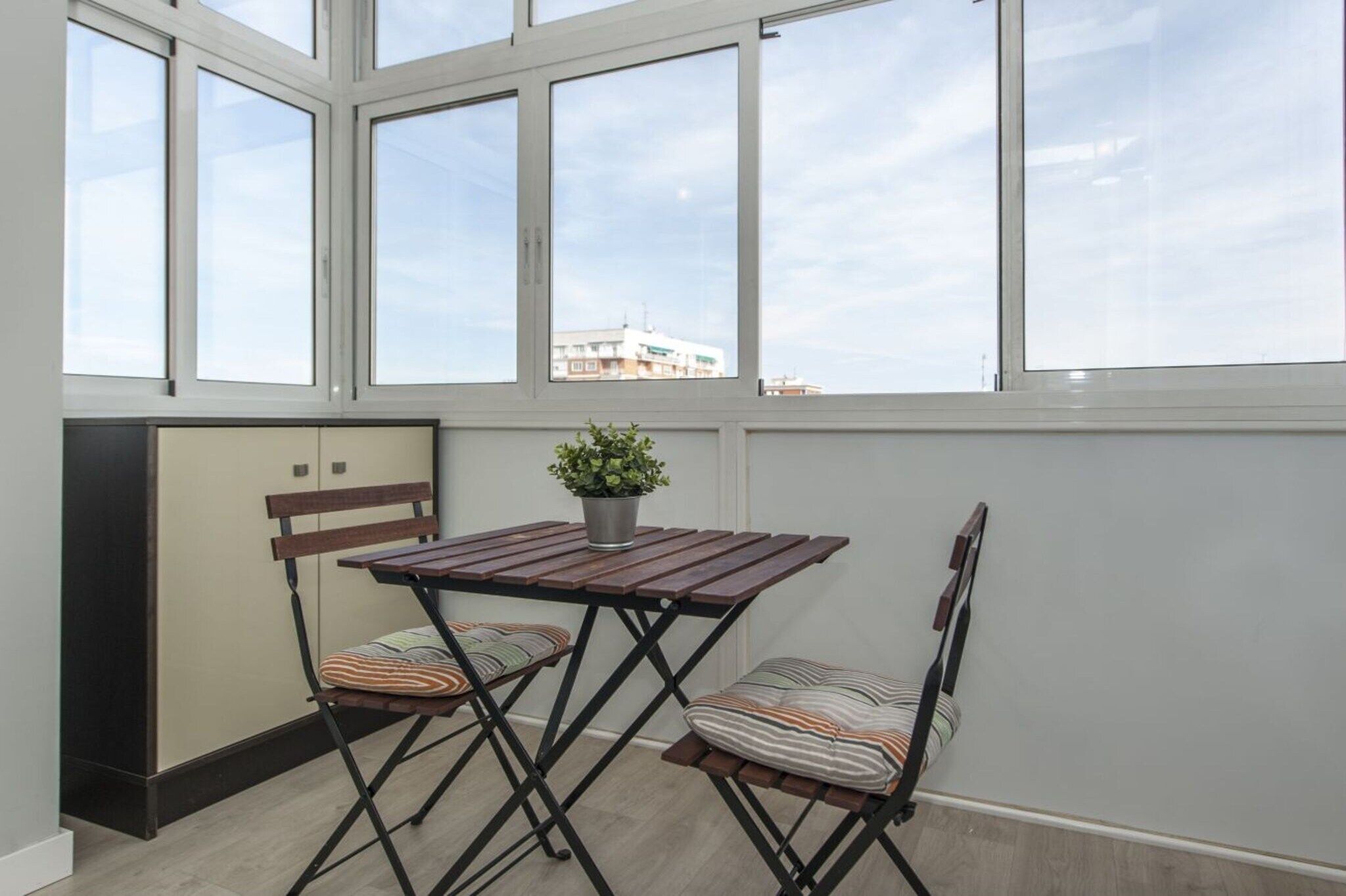 Une pièce avec une table et deux chaises, une plante en pot et une fenêtre donnant sur des immeubles.
