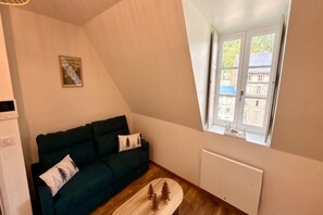A room with a dark green sofa, a small table with a decorative item, a radiator, and a window with a view of buildings. - Mont-Dore Apartment, Mountain View (Mont-Dore)