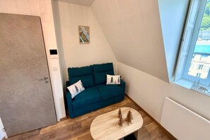 A small room with a teal sofa, a small wooden table, and a window with a view of buildings. - Mont-Dore Apartment, Mountain View (Mont-Dore)