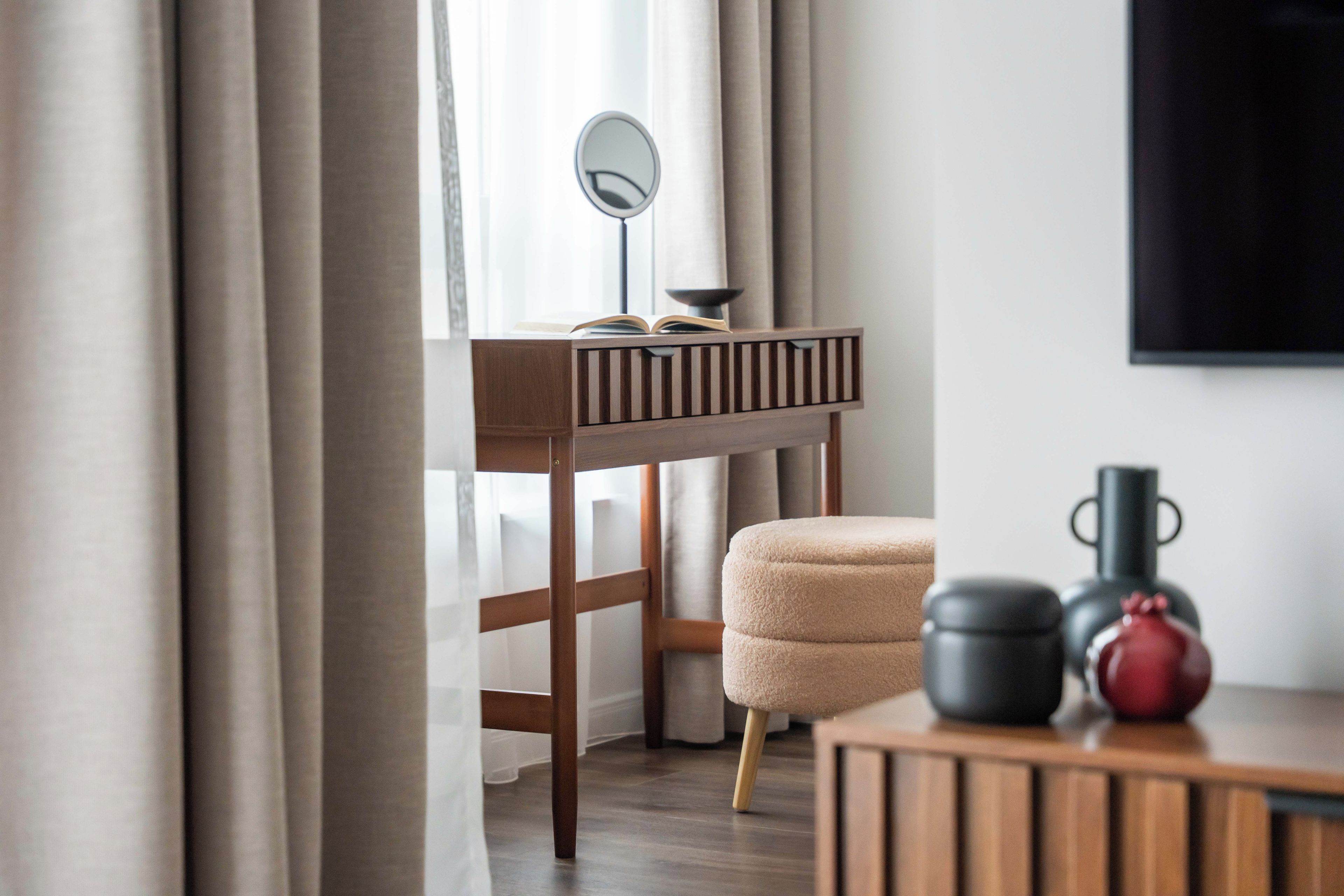 A room with a wooden desk, a round mirror, a beige chair, and a small table with a black vase and a red object.