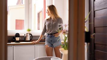 A person standing by a window with a view of buildings outside, a coffee maker, and a plant on the windowsill.