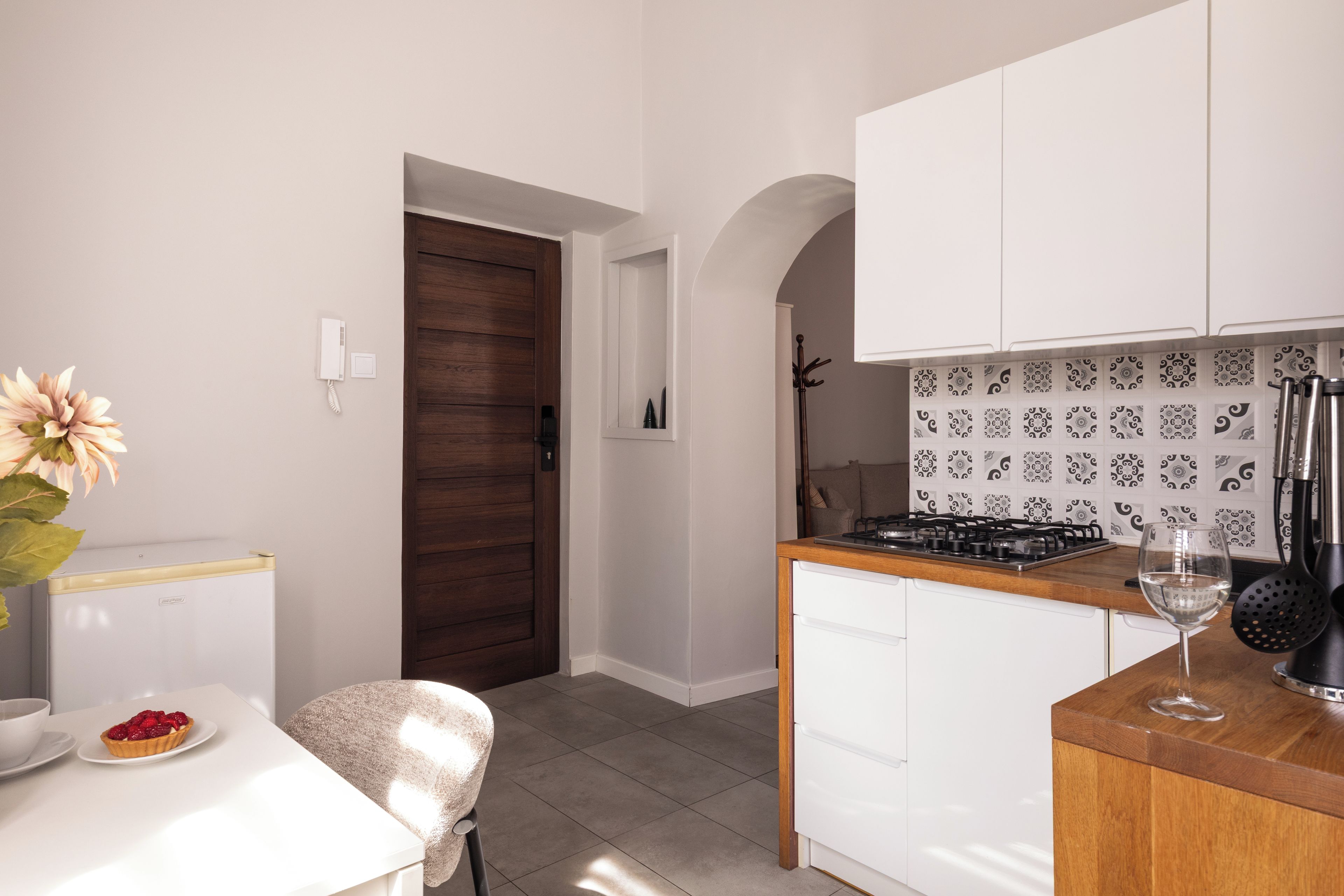 A compact kitchen with a white countertop, a stove, and a sink. There is a door leading to another room and a small table with a plate of food and a glass.