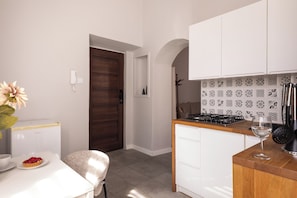 Una cocina compacta con encimera blanca, estufa y lavabo. Hay una puerta que da a otra habitación y una pequeña mesa con un plato de comida y un vaso.