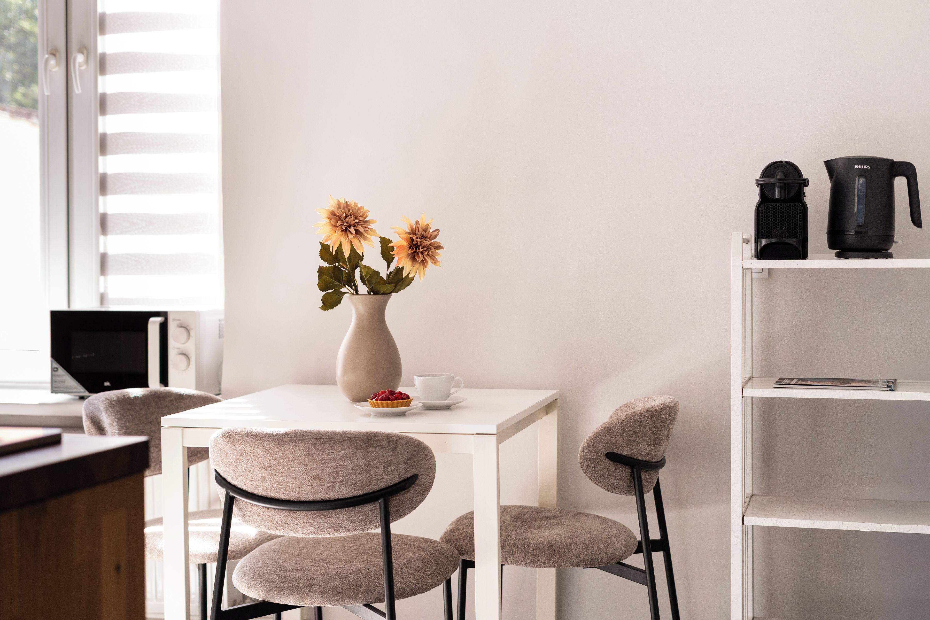 A minimalist room with a white table, two chairs, a vase with flowers, a coffee maker, and a bookshelf.