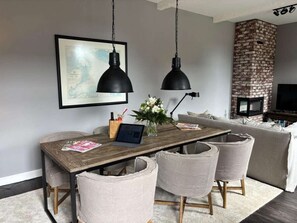 A modern living room with a wooden dining table, two black pendant lights, a framed map on the wall, a brick fireplace, and a grey sofa. - Strandhuus, Apartment 2 "strandlaufer" (Grossenbrode)