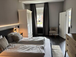 A bedroom with two beds, a wardrobe, a chair, and a view of the outside. - Strandhuus, Apartment 2 "strandlaufer" (Grossenbrode)
