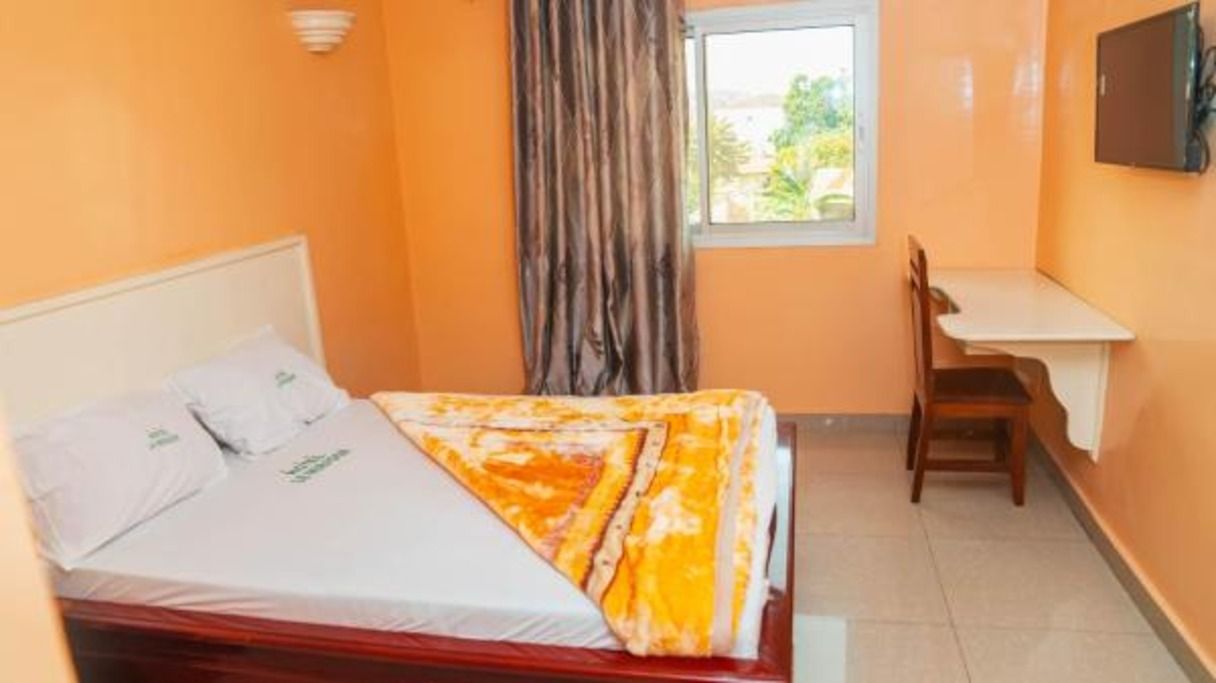 A single bed with a white mattress and pillows, a wooden headboard, a bedside table, a chair, a window with curtains, and a wall-mounted TV.