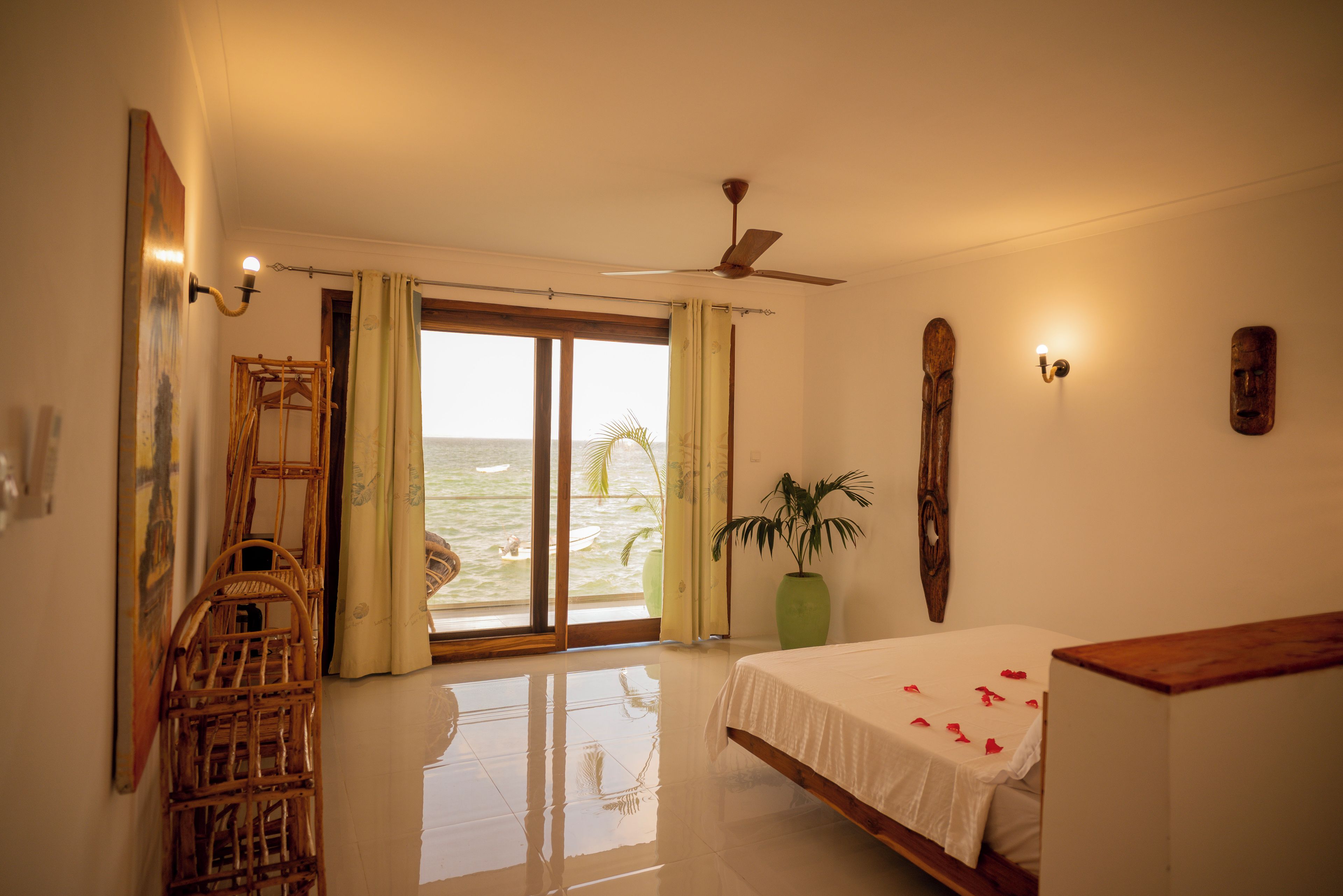 A bedroom with a bed, a wooden chair, a potted plant, a ceiling fan, and a view of the ocean through a sliding door.