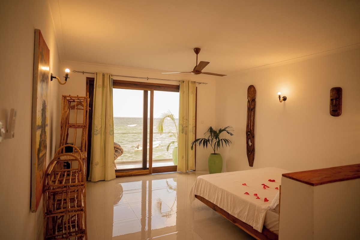 A bedroom with a bed, a wooden chair, a potted plant, a ceiling fan, and a view of the ocean through a sliding door.