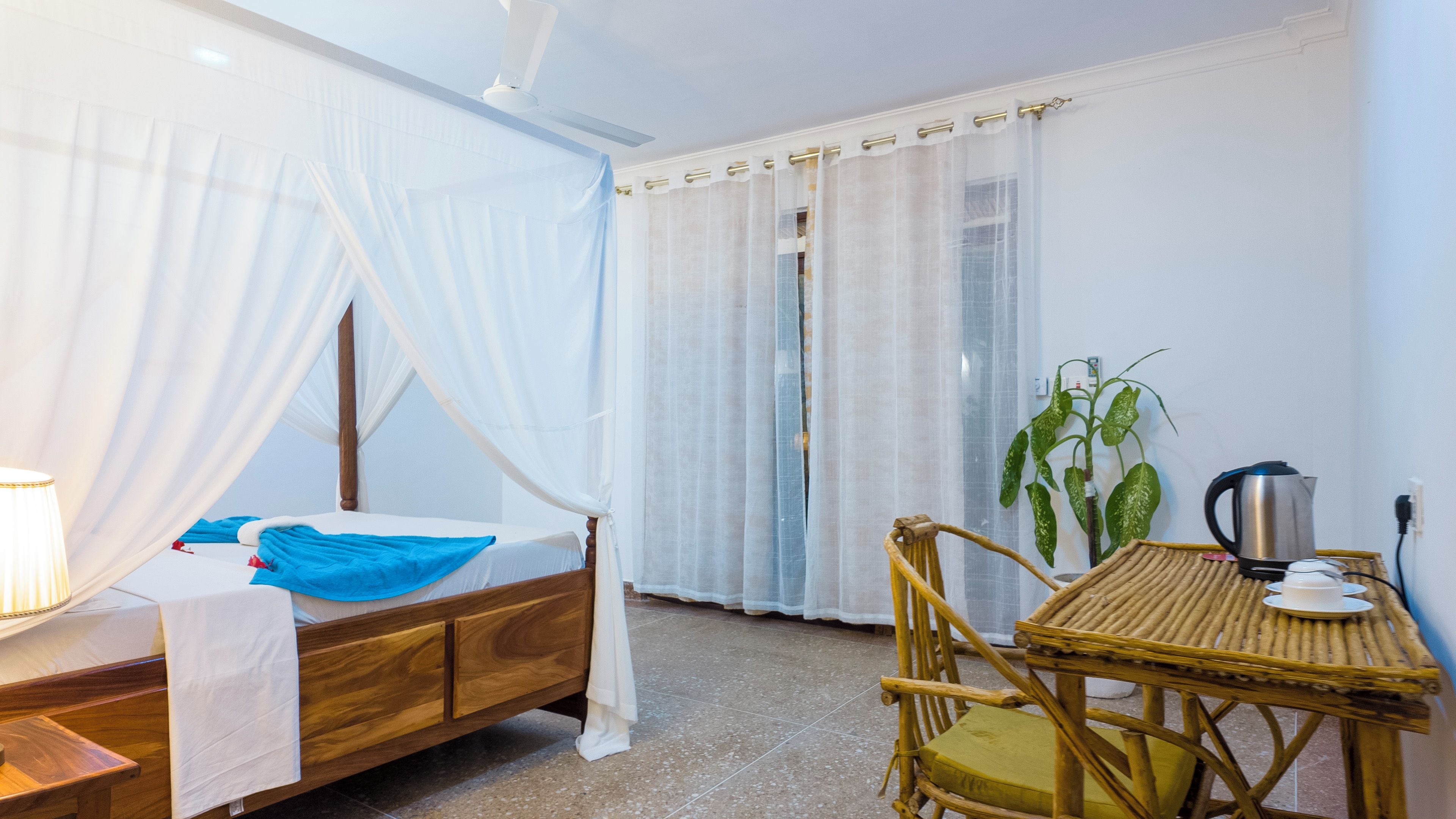 A room with a bed, a table with a kettle, a chair, and a plant.