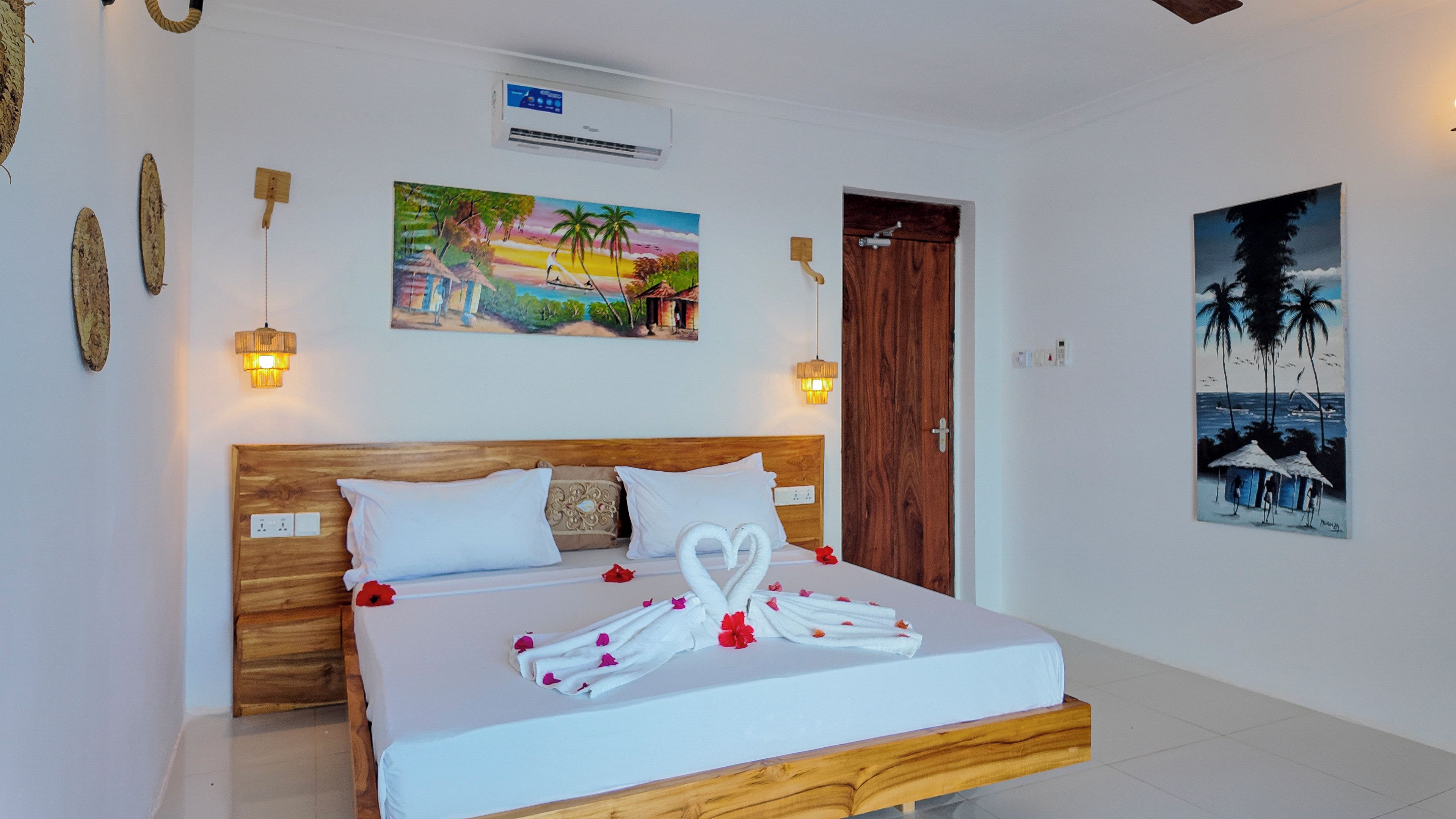 Une chambre d’hôtel avec un lit en bois, du linge de lit blanc, un tableau représentant un paysage tropical et une porte avec un cadre en bois.