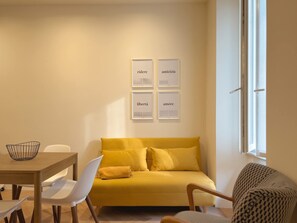 Een woonkamer met een gele bank, een eettafel met stoelen en een raam met jaloezieën.