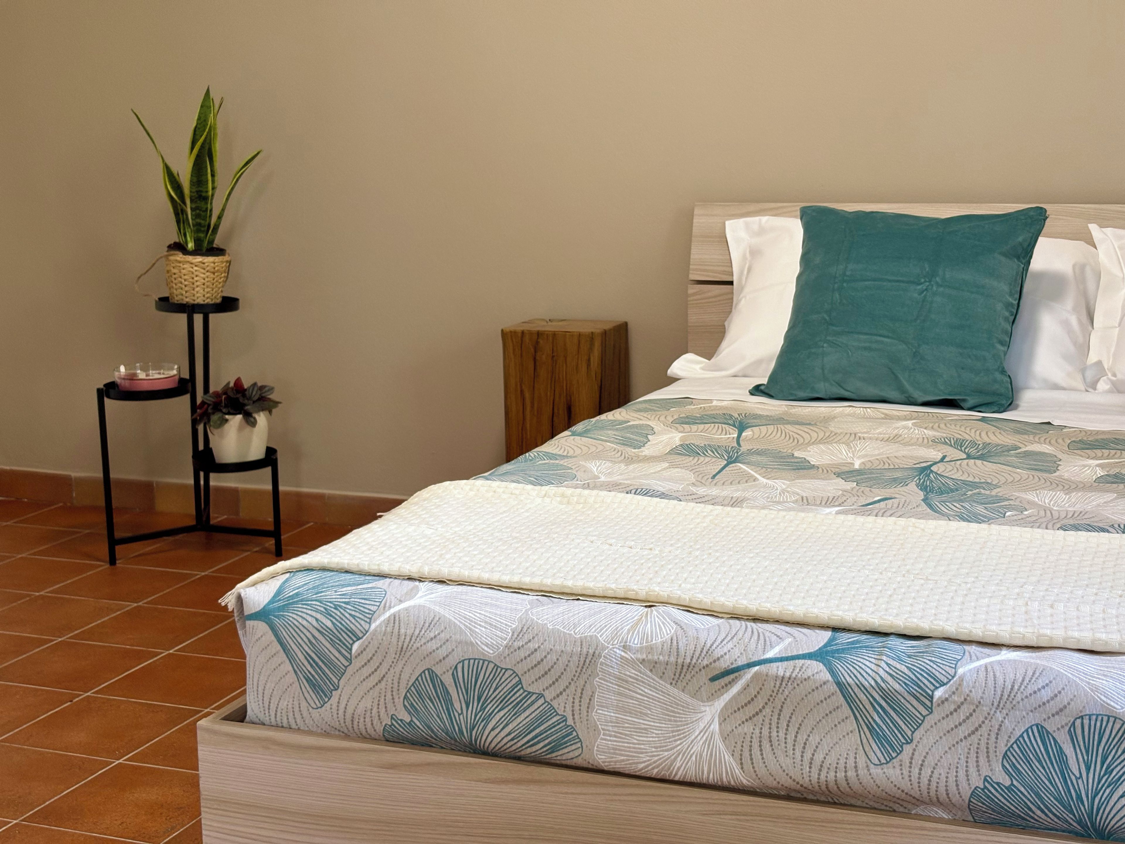 A neatly made bed with a floral-patterned bedspread, a wooden nightstand with a small potted plant, and a woven basket on a black stand.