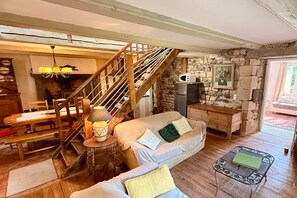 A cozy living room with a wooden staircase, a sofa with cushions, a dining table with chairs, and a kitchen area with a microwave. - Pondside cottage for six guests (Saint-Sauves-d´Auvergne)
