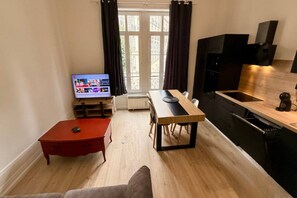 Un espace de vie compact comprenant une table à manger, une petite table d’appoint rouge, un canapé, un téléviseur et un coin cuisine avec une cuisinière et des placards.