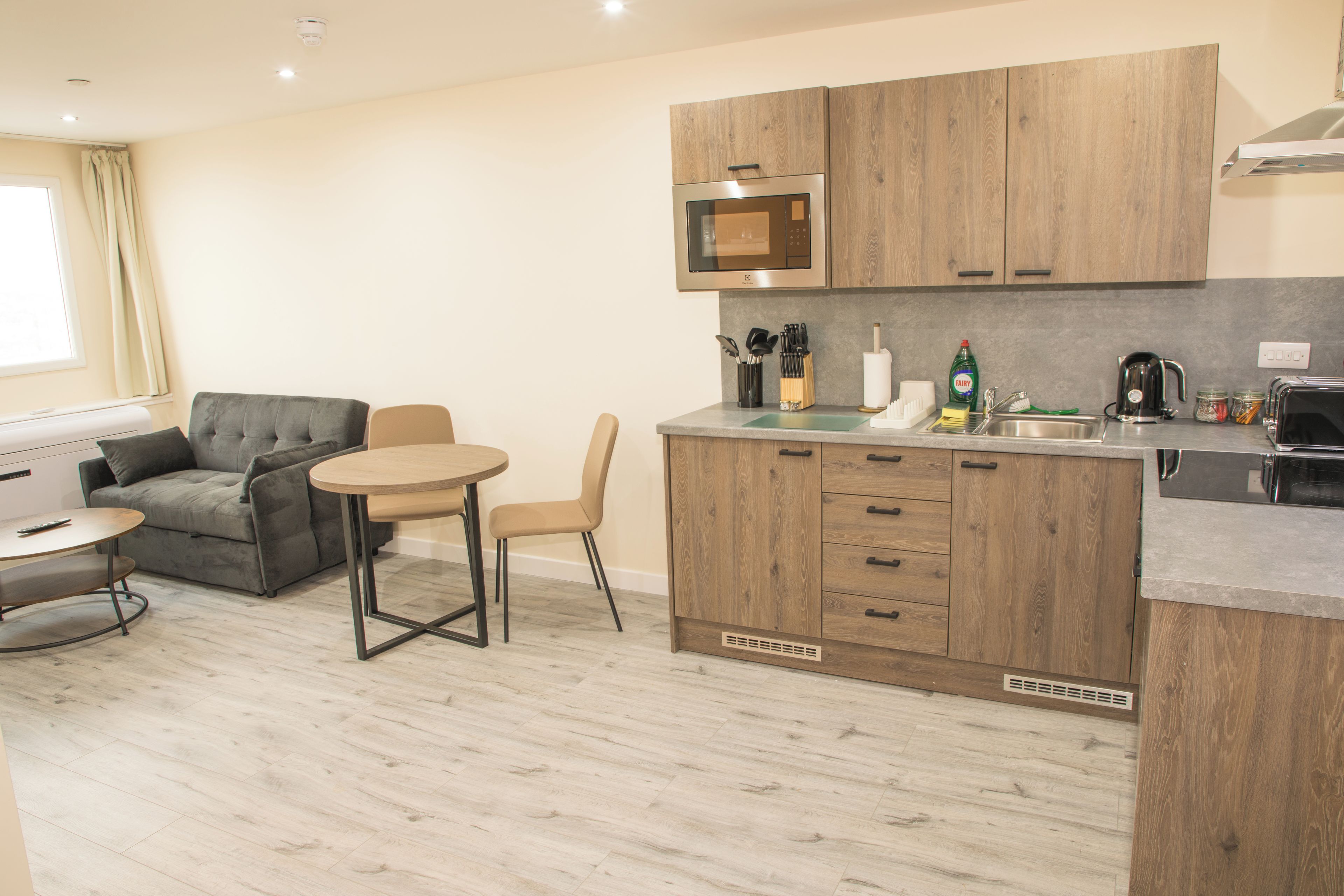 A compact kitchen with wooden cabinets, a microwave, and a coffee maker. There is a small dining area with a table and two chairs.