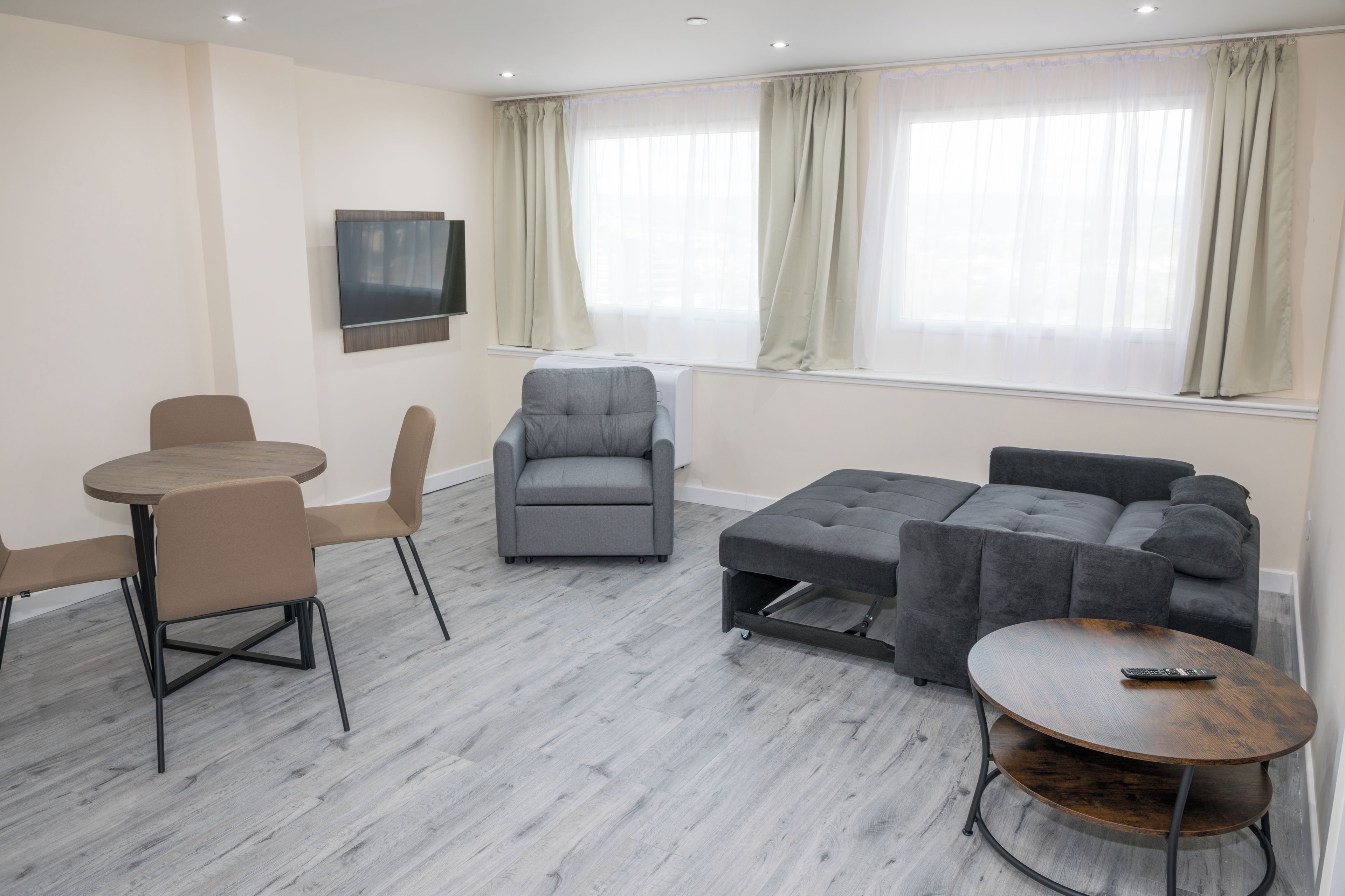A modern living room with a grey sofa, a grey armchair, a round wooden table, and a TV mounted on the wall.