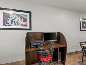 Ein Zimmer mit Schreibtisch, Drucker, Computermonitor und Stuhl. An der Wand hängen zwei gerahmte Bilder.