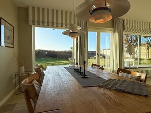 A dining area with a large wooden table, chairs, and a view of a grassy field through large windows. - Thatched House on the Beach (Wyk auf Foehr)