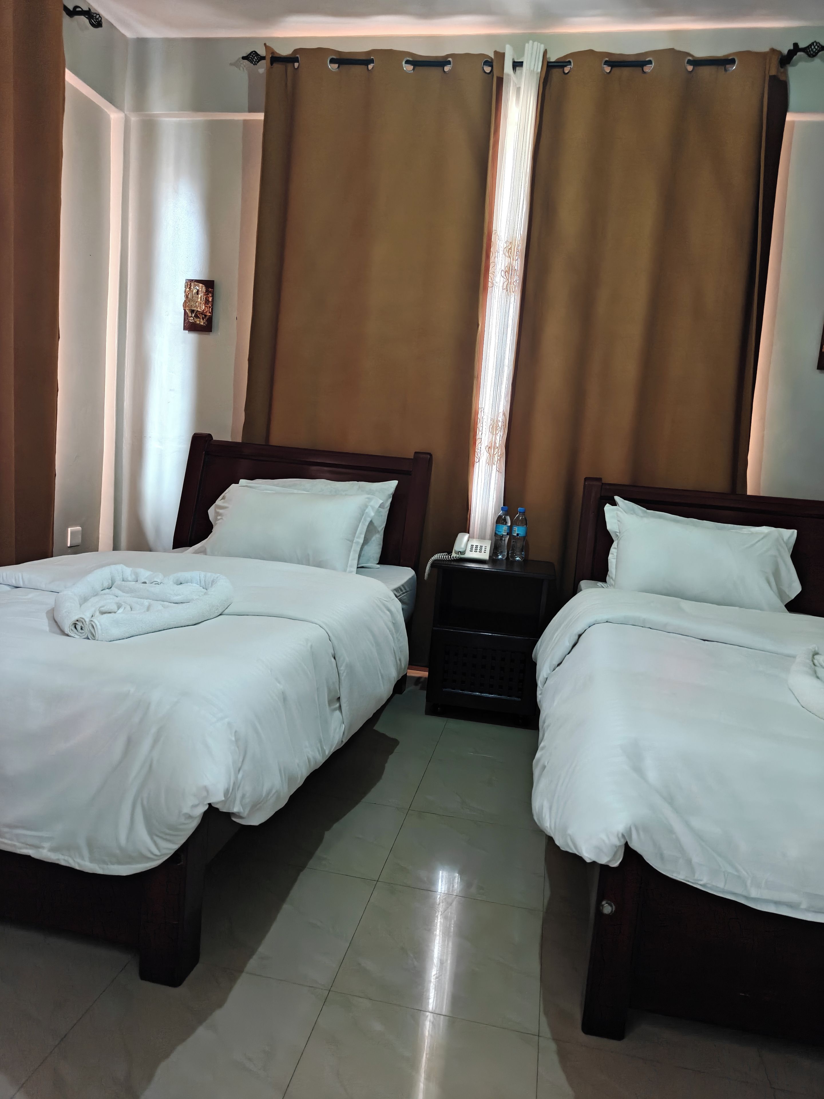 Ein Hotelzimmer mit zwei Betten, einem Nachttisch mit Telefon und Wasserflaschen sowie einem Vorhang.