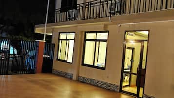 Un edificio de varios pisos con techo plano, entrada protegida por una reja y un interior bien iluminado visible a través de las ventanas.