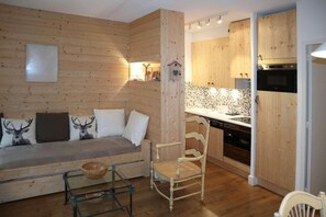 A cozy cabin interior with a wooden wall, a sofa with deer-themed pillows, a dining table with chairs, and a kitchen area with wooden cabinets and appliances. - 1st floor, balcony, parking, tv, ski locker, 55m², Les Orres (Les Orres)
