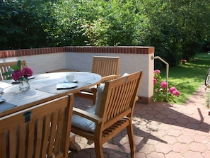 An outdoor patio with a round table and wooden chairs, surrounded by a brick wall and a garden with pink flowers. - "holidays in a Former Captain's House" (Wyk auf Foehr)