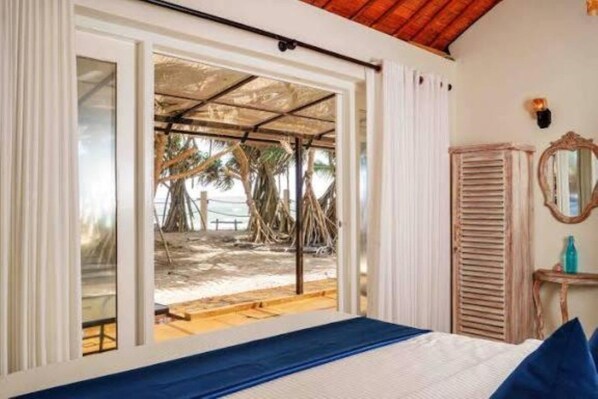 A room with a bed, a window with a view of a beach, and a wooden ceiling.